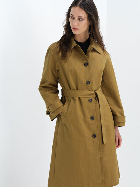A woman wearing a khaki longline belted trench coat with long sleeves, a pointed collar and button down fastening.