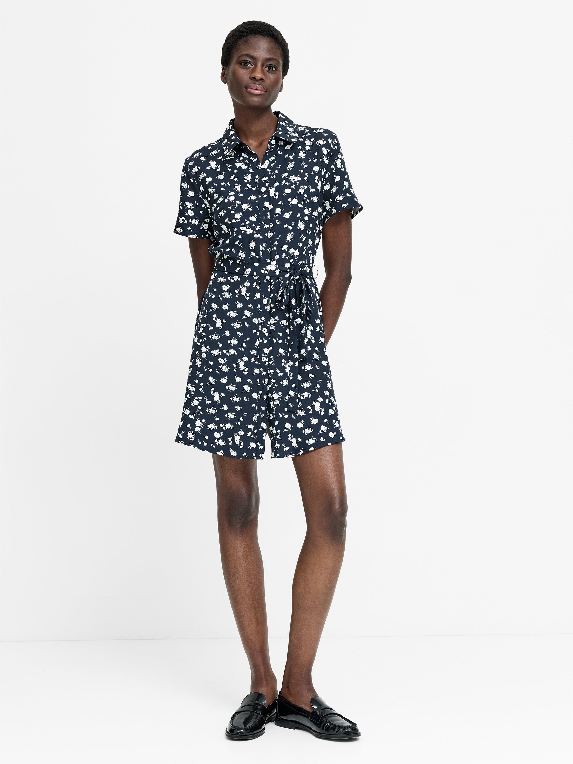A woman wearing a short sleeve mini length navy dress with a white floral print, shirt collar, self tie belt and a button down fastening. 