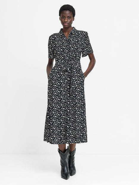 A woman wearing a short sleeved black midi length dress with a shirt collar, button down fastening, self-tie drawstring waist, two pockets and a white floral pattern.
