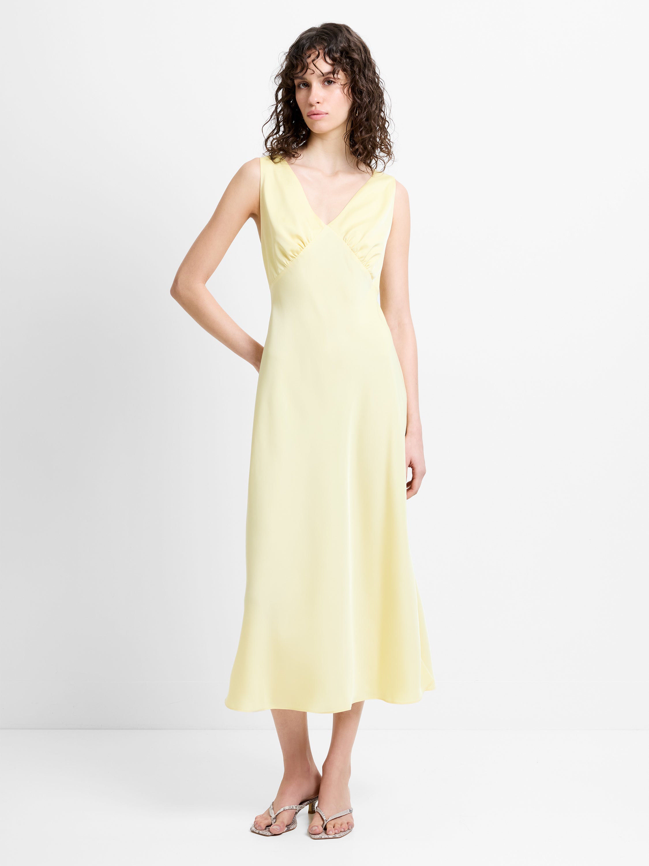 Woman wearing a pale yellow sleeveless satin midi dress, with a V neckline and ruching waist detailing