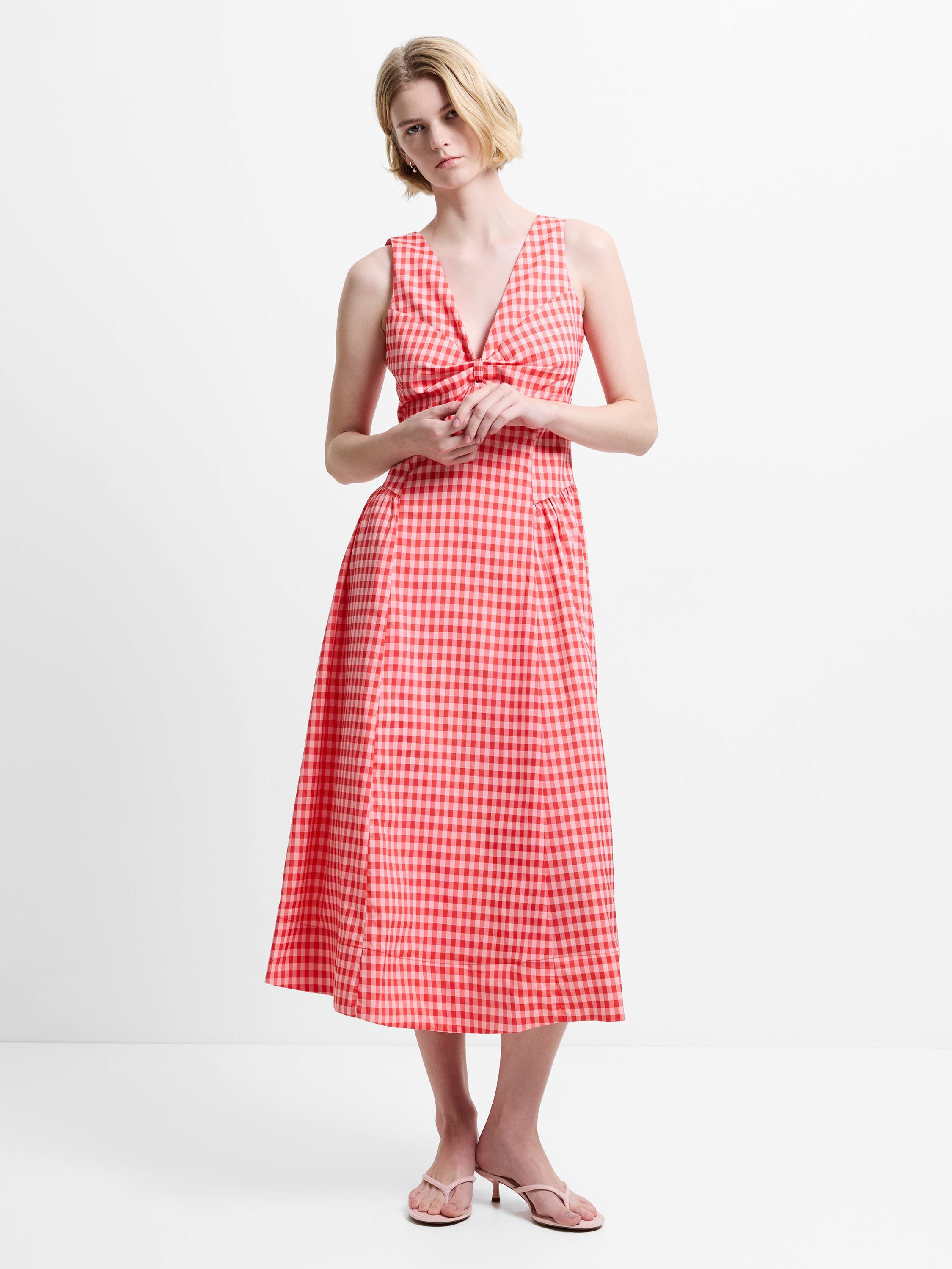 Woman wearing a red gingham sleeveless midi dress, with a deep V neckline and an empire waist with a twist knot detail
