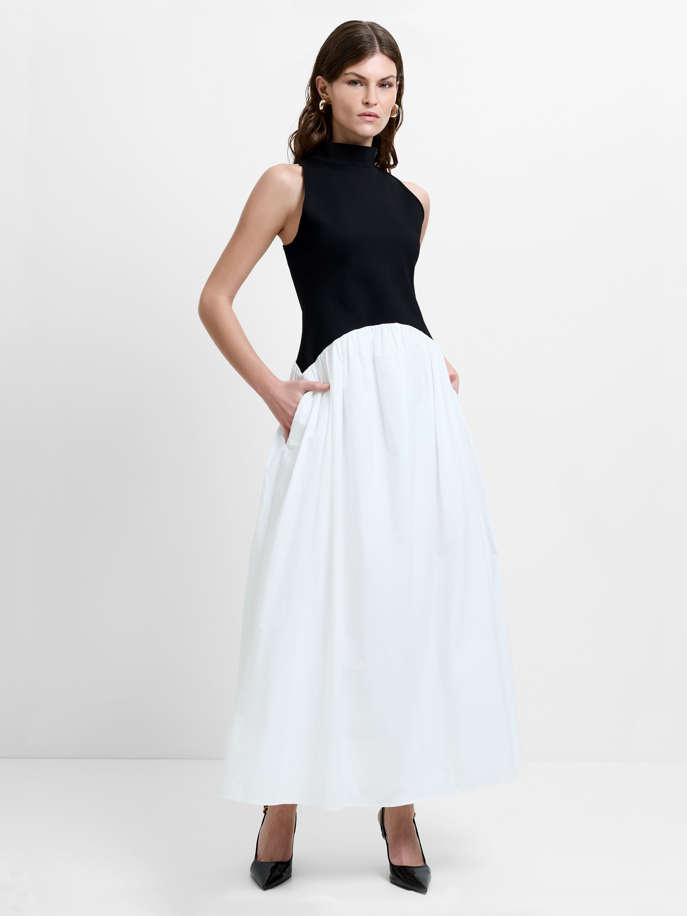 Woman wearing a sleeveless colour block design maxi dress, with a black high neck bodice and a white voluminous pleated skirt