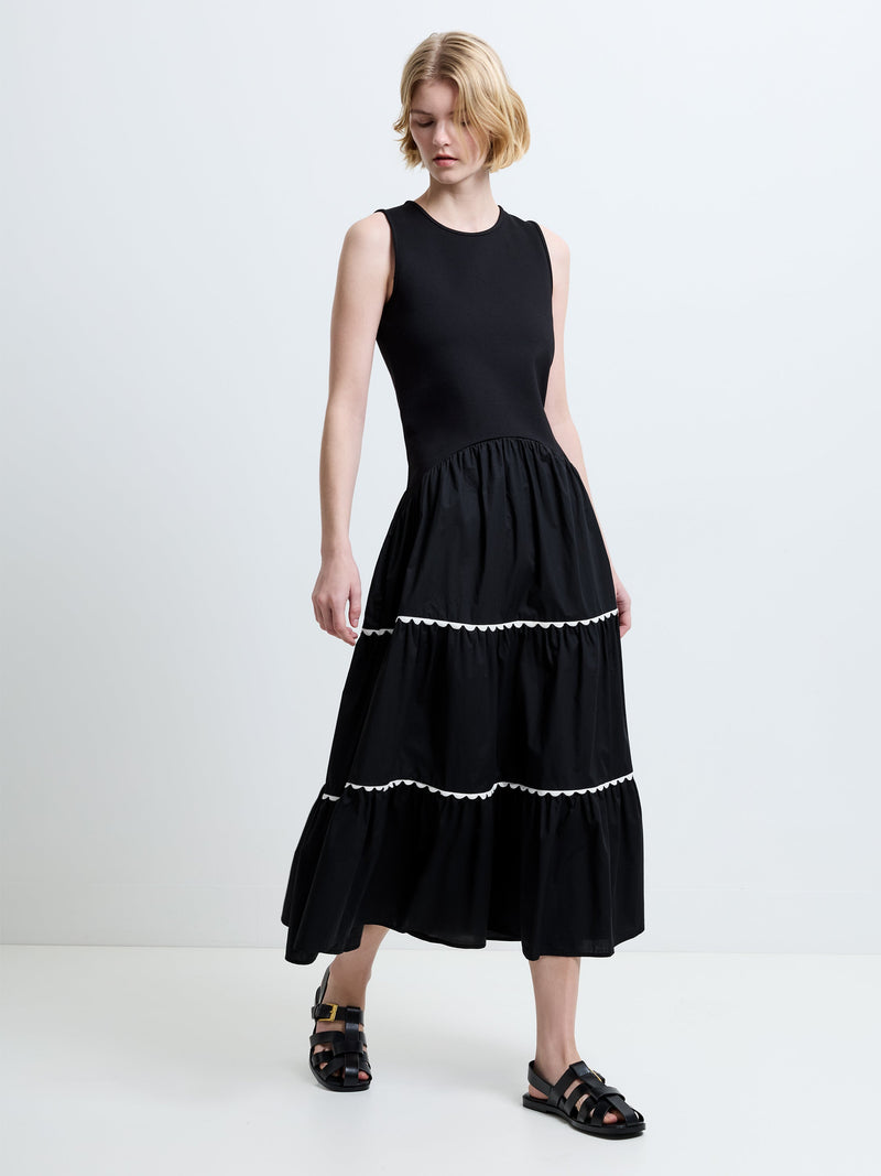 Woman wearing a black dropped waist sleeveless midi dress, with a round neckline and ricrac white trims on the skirt