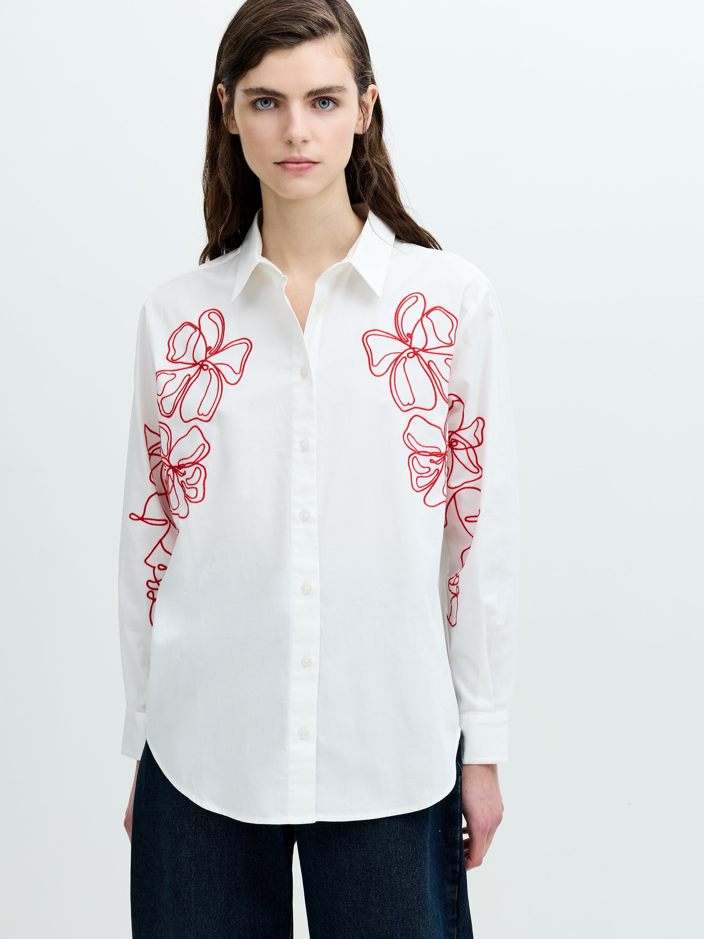 A woman wearing a white long sleeved shirt with red floral embroidery, a pointed collar and a back box pleat.