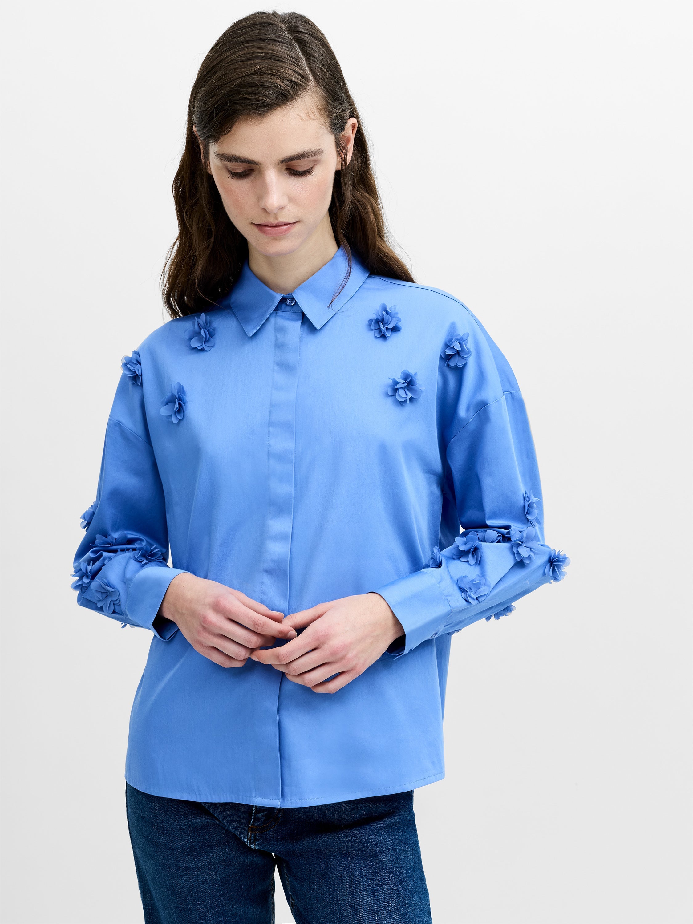 A woman wearing a blue cotton shirt with a 3D floral print, long sleeves, button down front, shirt collar, pleated back, dropped shoulders and a relaxed boxy fit.