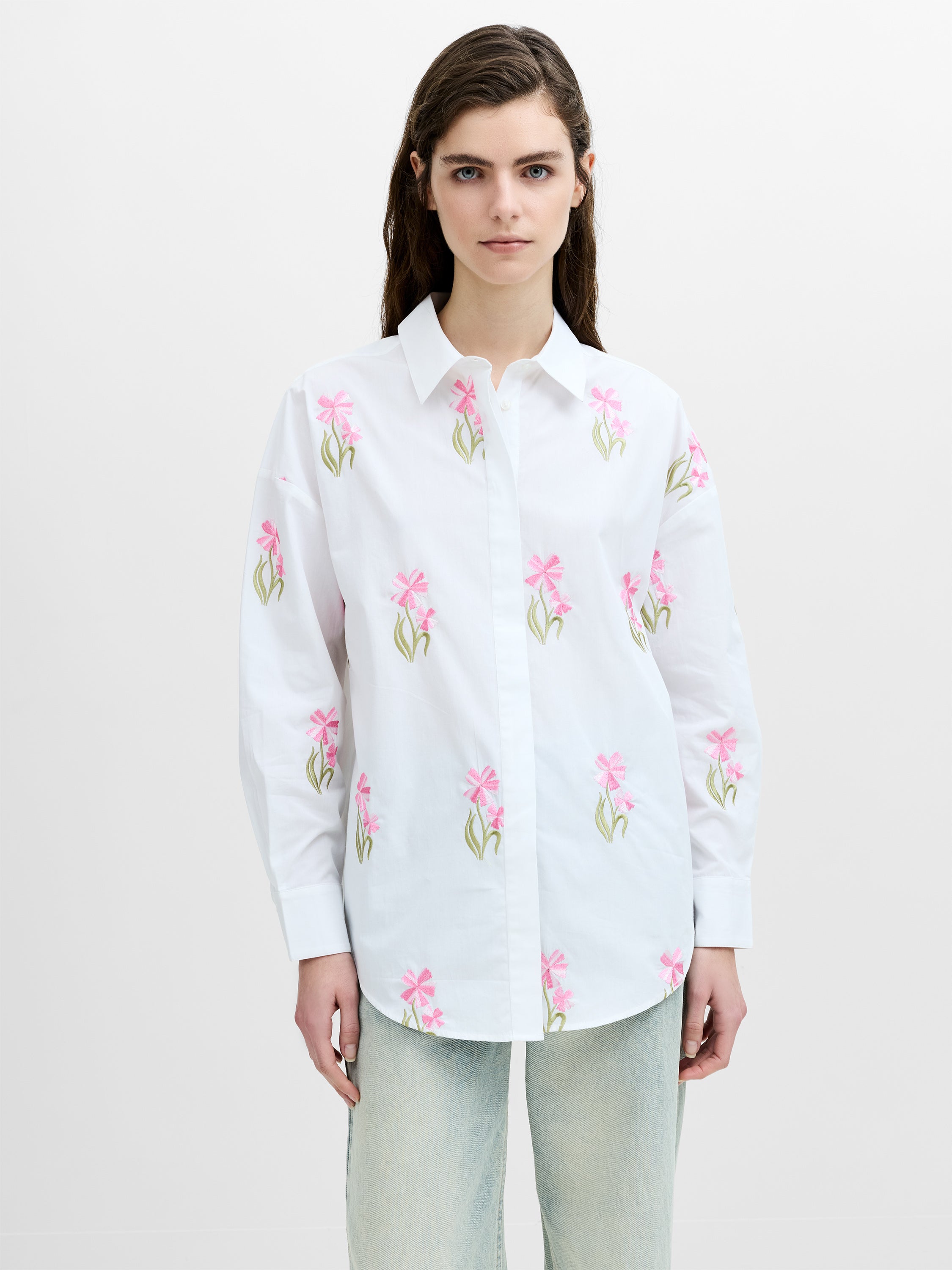 A woman wearing a cotton white shirt with a pink and green floral embroidered front, point collar, button down fastening and long sleeves.