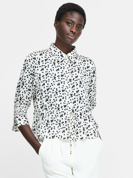 A woman wearing a cream button down shirt with a black floral pattern, 3/4 length sleeves, a collared neckline, and a self tie drawstring waist.