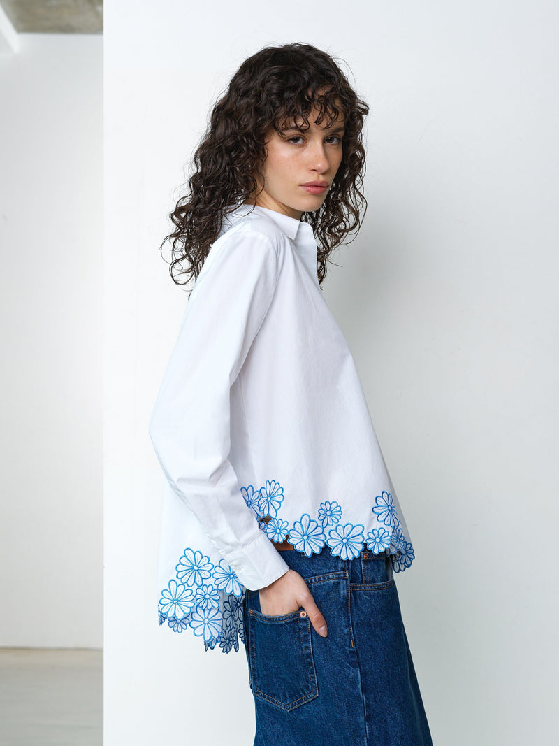 Woman wearing a long sleeve white shirt with blue floral cutwork embroidered hem detailing