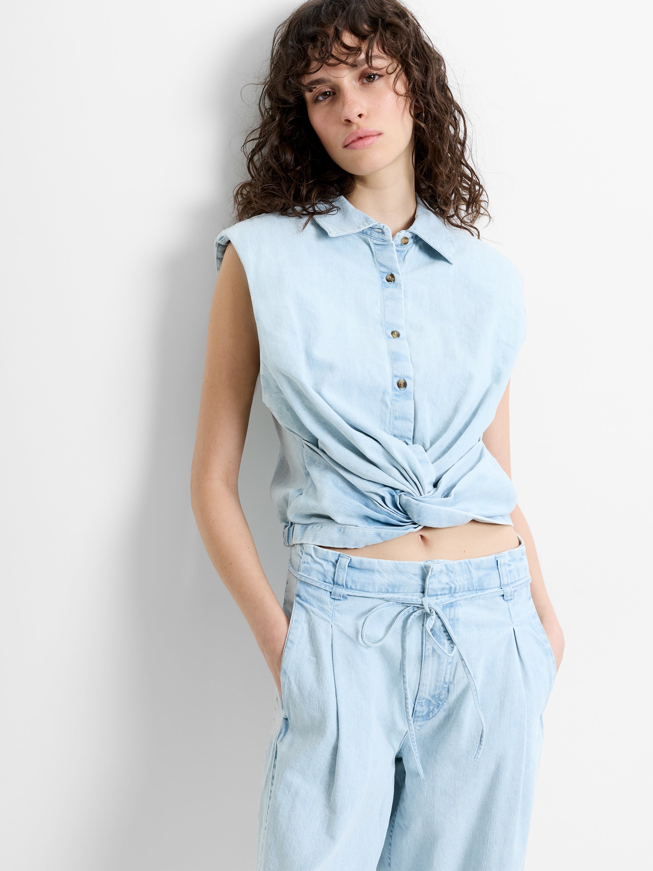 Woman wearing a light blue denim sleeveless top, with padded shoulders and a front twist knot detail