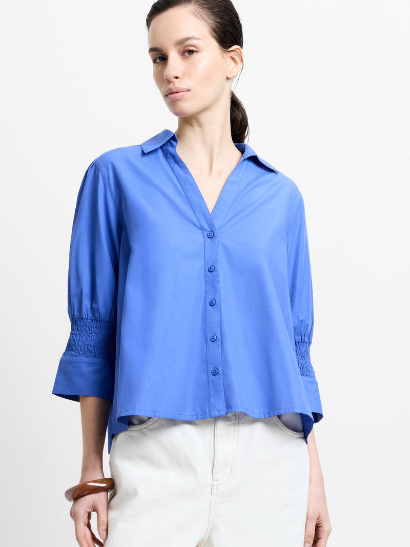 Woman wearing a blue V-neck shirt with 3/4 length sleeves with smocked cuffs and pleated tie back detailing