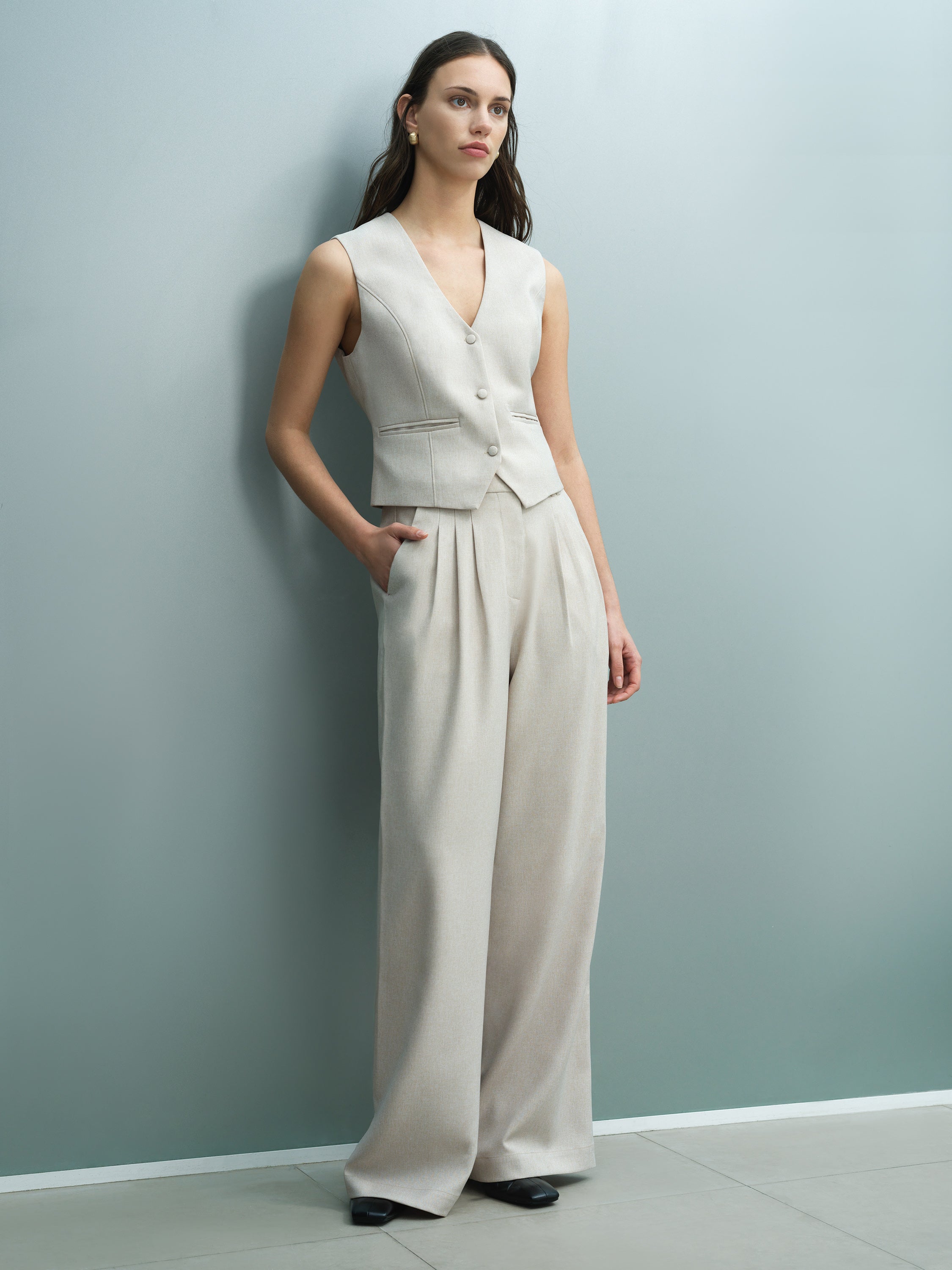 A woman wearing tailored high waisted, full length pleated, wide leg, beige trousers with four pockets.