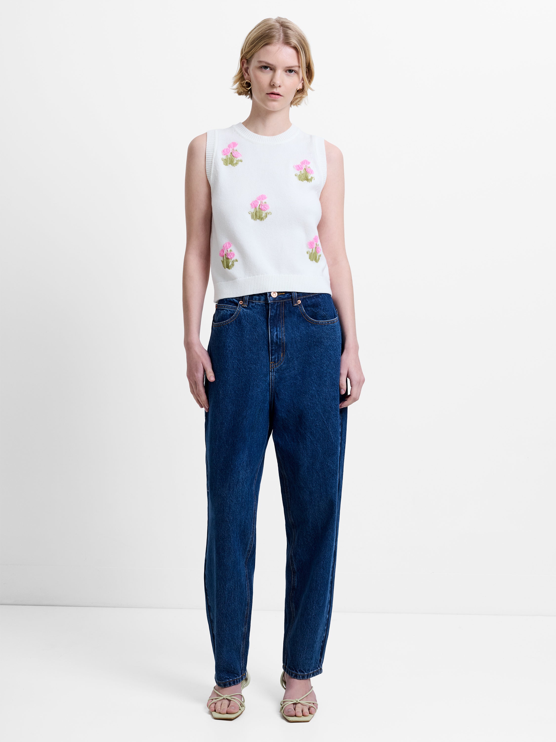 Woman wearing a cream knitted vest top with floral embellishments and dark blue mom barrel leg jeans