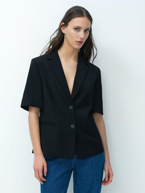 A woman wearing a tailored black short sleeved blazer with a collared V-neckline and 2 front buttons. 