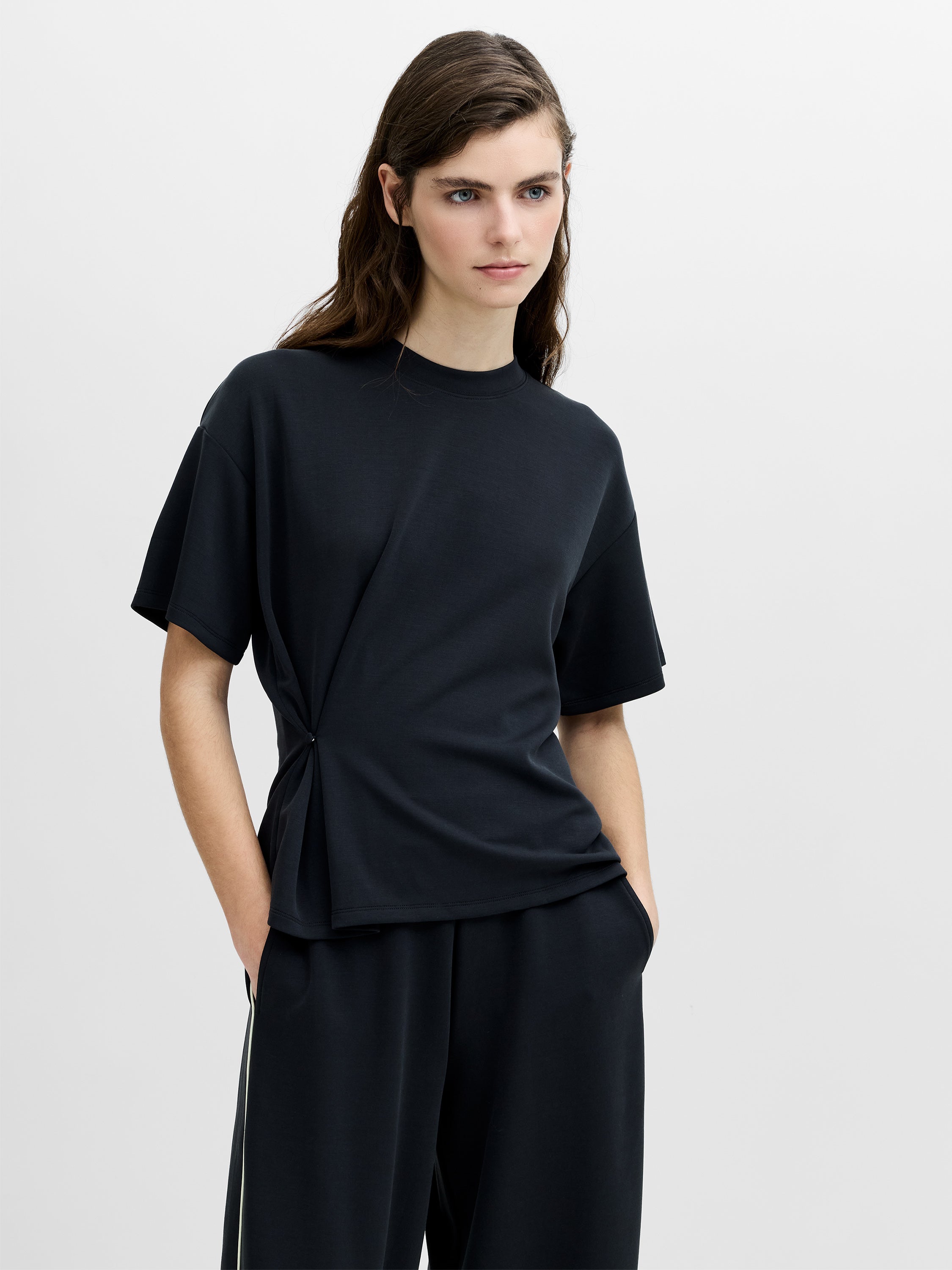 A woman wearing a dark grey relaxed fit T-shirt with a cinched waist, dropped shoulders, a crew neck and short sleeves.