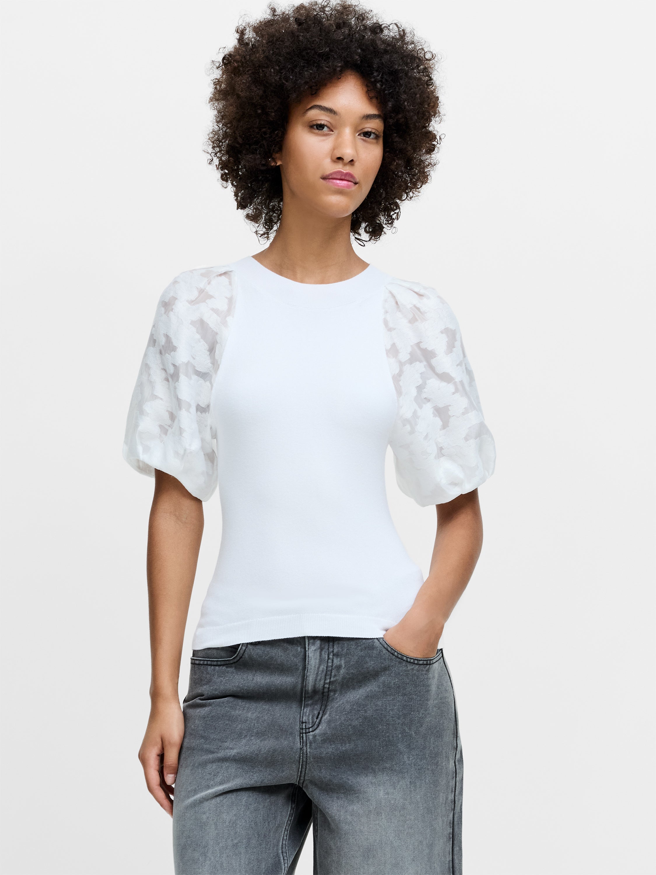 A woman wearing a white knit top with sheer puff sleeves, a crew neckline and a ribbed hem.