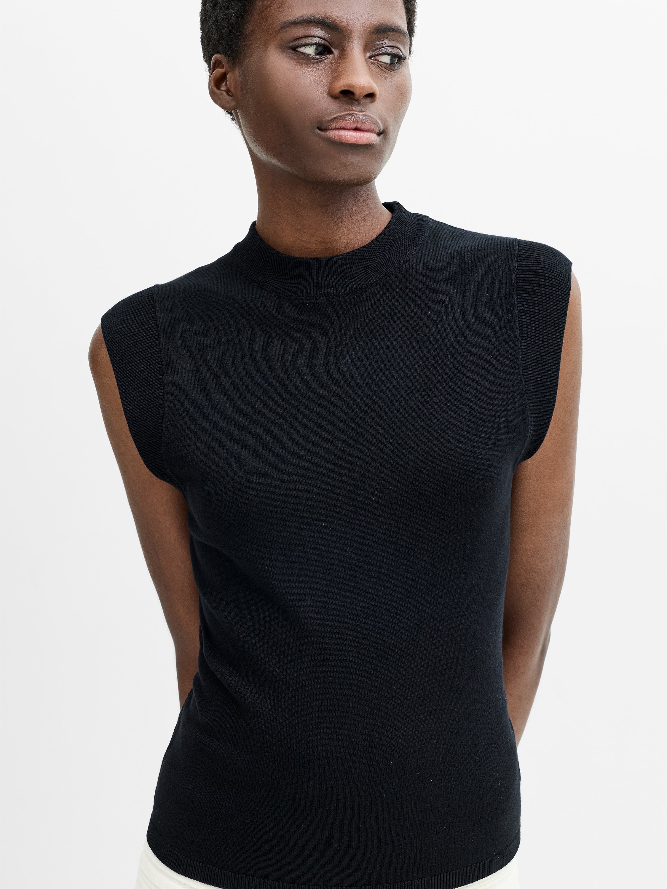 A woman wearing a black fitted, knit sleeveless top with an adjustable tie back and mock neckline. 