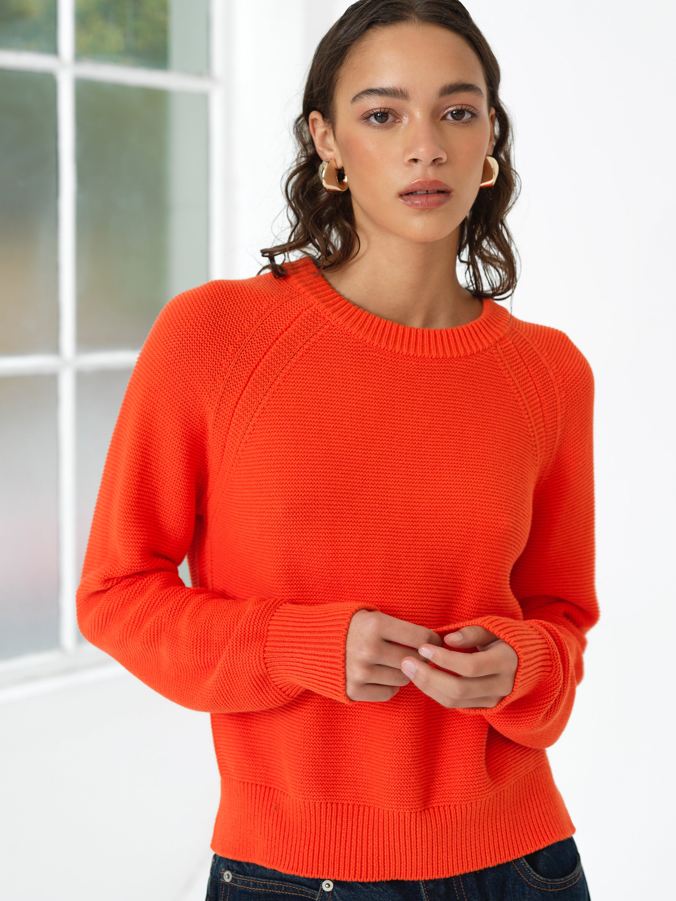 Woman wearing a bright orange crew neck knitted jumper with long raglan sleeves and a ribbed crew neck and hem