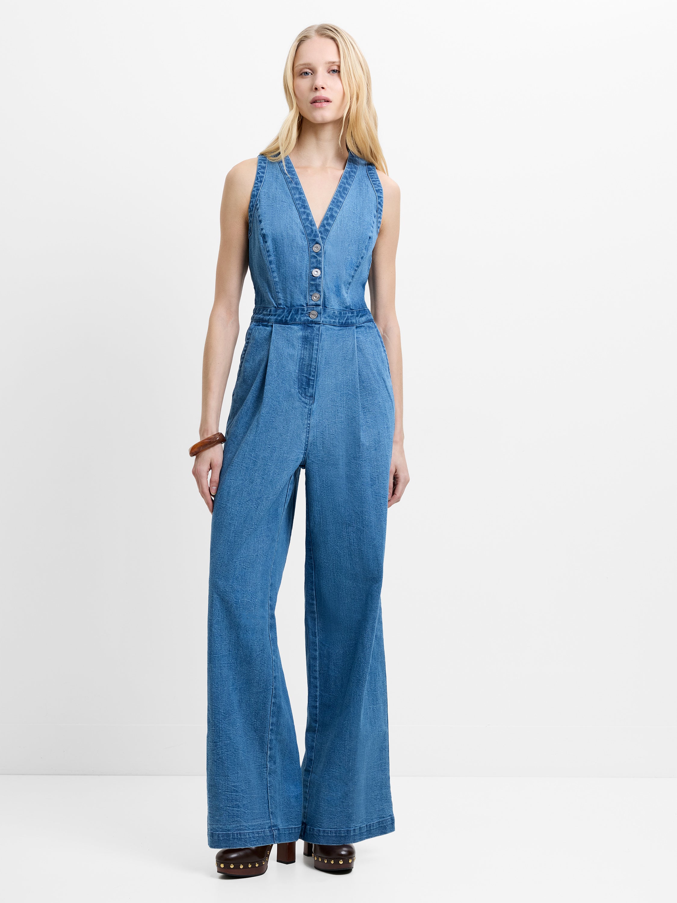 Woman wearing a blue denim sleeveless wide leg jumpsuit with a V neckline and metal button fastening